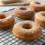 Four golden-brown cinnamon sugar donuts resting on a black cooling rack, lightly dusted with cinnamon sugar, with a soft and fluffy texture. A wooden tray and scattered sugar granules add a cozy background."