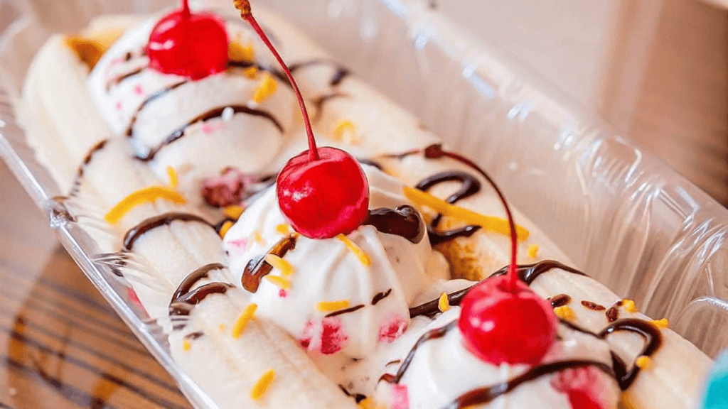 "A classic banana split served in a glass dish with scoops of chocolate, vanilla, and strawberry ice cream, each topped with whipped cream and cherries, with a fresh banana split lengthwise underneath."