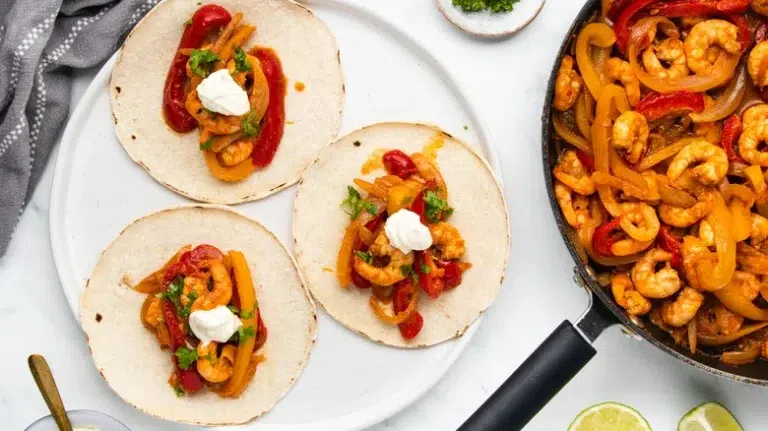 "Plate of shrimp fajitas with colorful bell peppers, onions, and lime wedges, served on a tortilla and topped with sour cream, alongside a cast-iron skillet with more fajita filling."