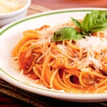 "A plate of spaghetti in tomato basil sauce garnished with fresh basil leaves and Parmesan cheese, served on a rustic wooden table with tomatoes, garlic, and olive oil in the background."