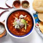 "A steaming bowl of pozole rojo garnished with shredded cabbage, sliced radishes, lime wedges, and oregano, served with tostadas on the side."