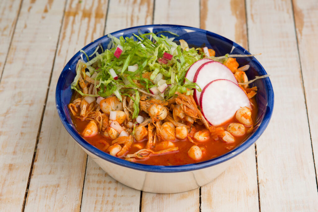 "A steaming bowl of pozole rojo garnished with shredded cabbage, sliced radishes, lime wedges, and oregano, served with tostadas on the side."