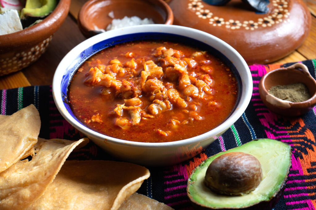"A steaming bowl of pozole rojo garnished with shredded cabbage, sliced radishes, lime wedges, and oregano, served with tostadas on the side."