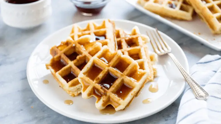 Golden brown Belgian waffles with deep pockets, drizzled with maple syrup, topped with a pat of butter, and lightly dusted with powdered sugar on a white plate.