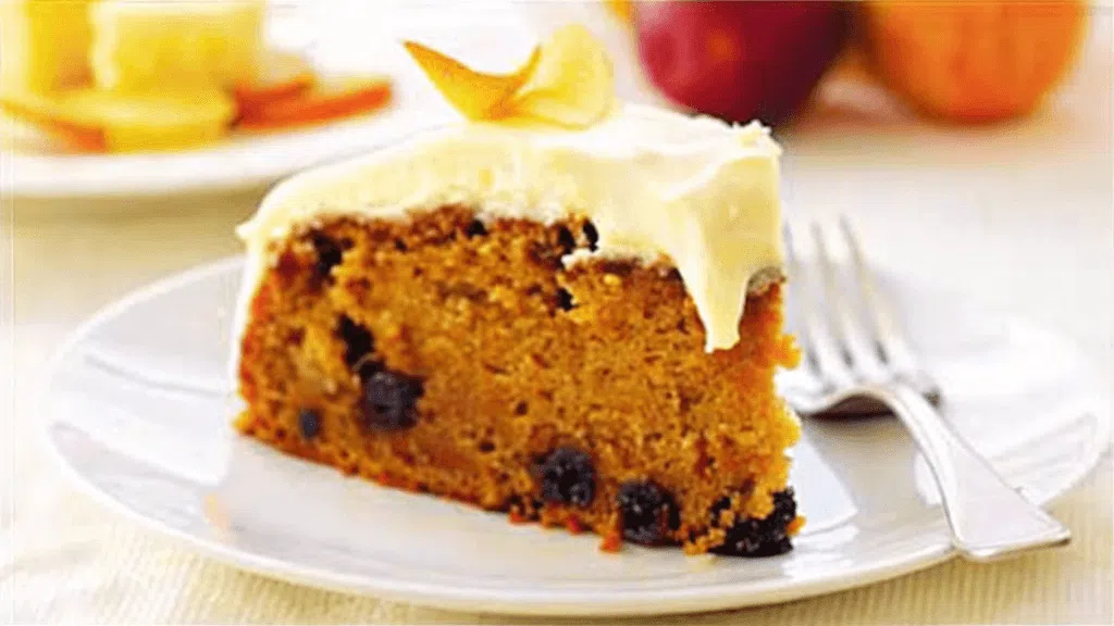 A close-up of a slice of layered sugar-free carrot cake with creamy frosting and topped with pecans, served on a white plate.