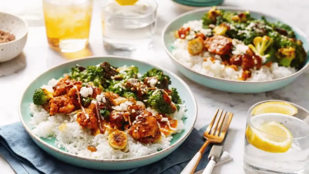 “Two bowls of sticky chicken rice with broccoli, drizzled with sauce, served over white rice, next to lemon water and iced drinks on a marble table.”