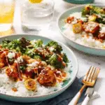 “Two bowls of sticky chicken rice with broccoli, drizzled with sauce, served over white rice, next to lemon water and iced drinks on a marble table.”